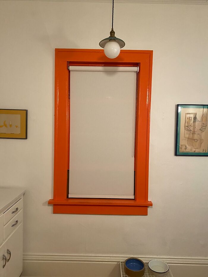 Orange window frame with a white roller blind, pendant light above, and framed wall art, a unique decor piece for magical homes.