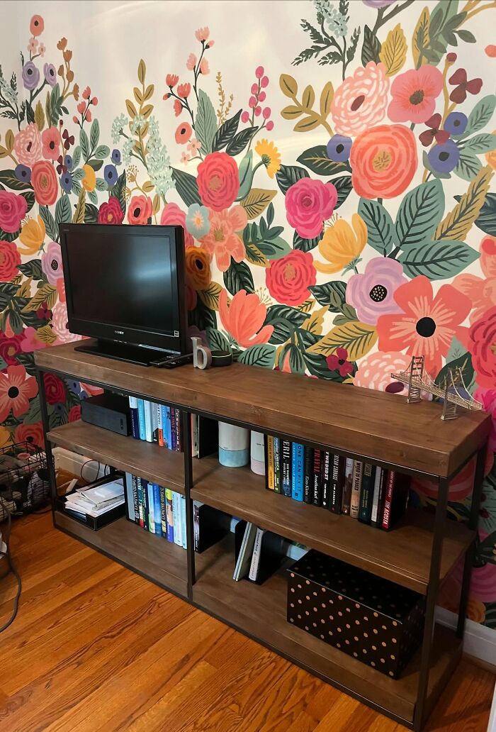 Wooden bookshelf with books and a TV in front of a vibrant floral wallpaper decor piece in a cozy home setting.