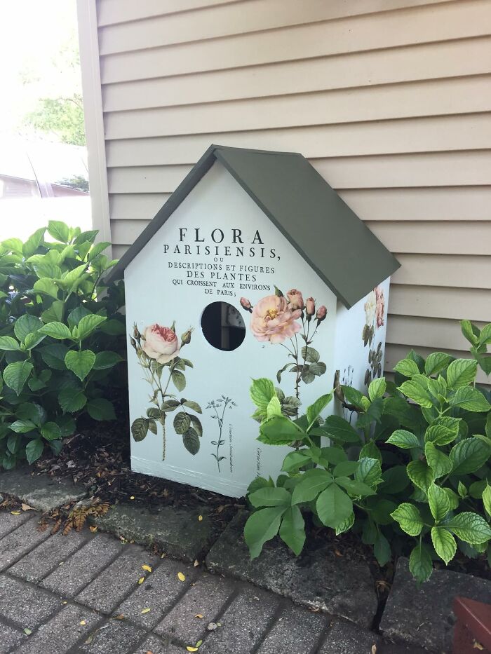 Decor piece with floral design and French text placed among green plants along a home exterior wall.