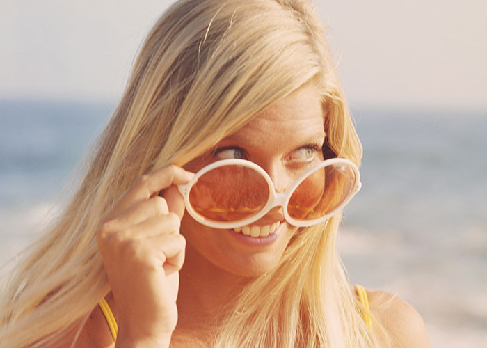 Blonde woman at the beach looking over oversized sunglasses, capturing a moment of unexplained mystery.