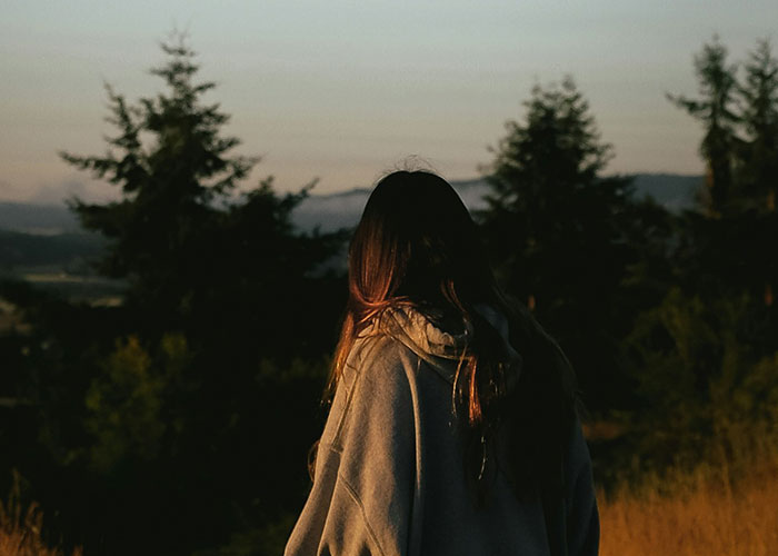 Woman with long hair wearing a hoodie, standing outdoors at dusk surrounded by trees, evoking mysteries people experienced.