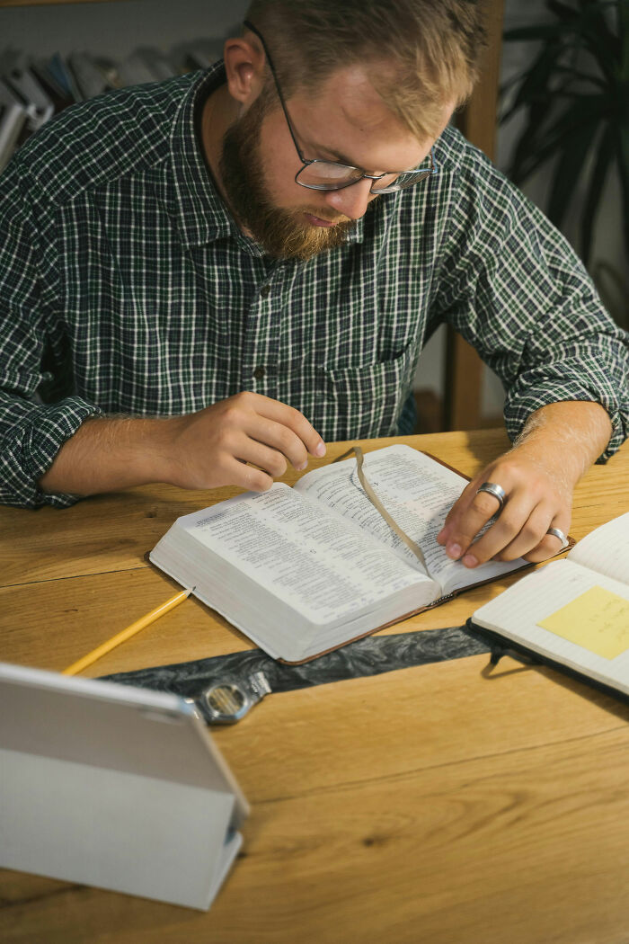 Man with glasses reading a book at a wooden table, focusing on study with flex accessories nearby and notes on the side