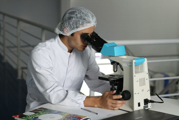 Scientist in lab coat and hairnet examining samples with a microscope, representing disturbing facts research.
