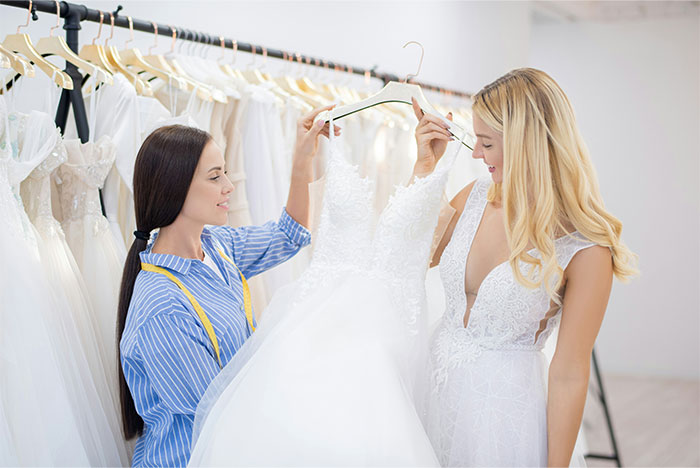 Two women in a bridal shop, one overworked and underpaid, looking at a white wedding dress on a hanger.
