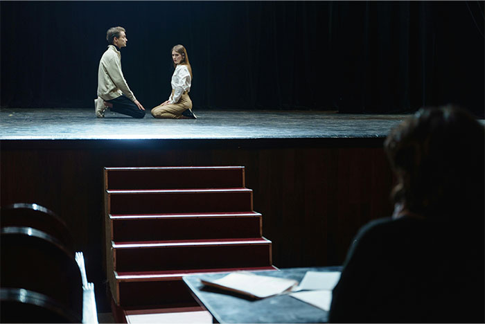 Two actors kneeling on a dimly lit stage during an audition at a struggling, overworked, and underpaid company.