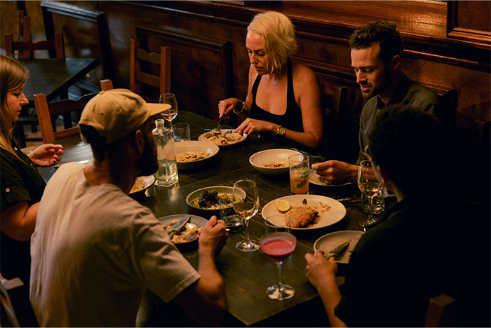 Group of overworked and underpaid employees sharing a meal at a dimly lit wooden table in a casual setting.