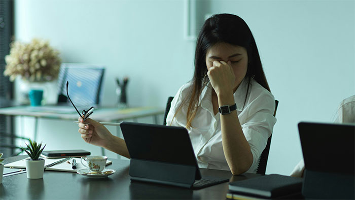 Tired woman at desk holding glasses, stressed from being overworked and underpaid in a struggling company environment.