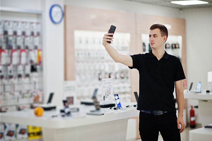 Young man in black shirt taking a selfie inside a retail store, illustrating overworked and underpaid employees at dying companies.