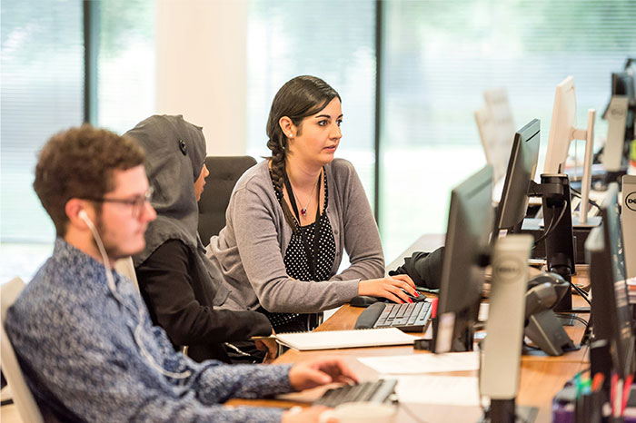 Three diverse employees working at computers in an office, illustrating overworked and underpaid company conditions.