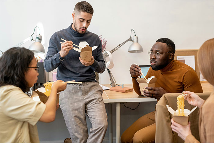 Young diverse coworkers eating takeout noodles together during a break, illustrating overworked and underpaid office culture.