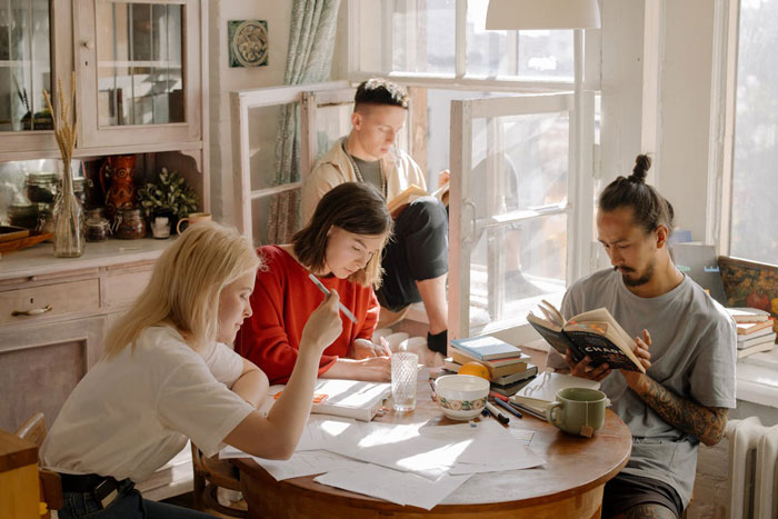 Nonbinary person sitting with roommates in a sunlit room, reading and writing at a wooden table with books and papers. Nonbinary person sitting with roommates in a sunlit room, reading and writing at a wooden table with books and papers.