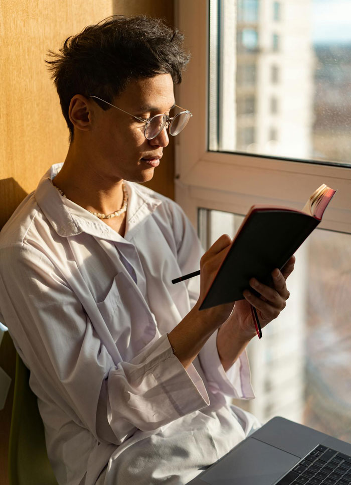 Nonbinary person wearing glasses sitting by window, writing in notebook with laptop nearby in a sunlit room. Nonbinary person wearing glasses sitting by window, writing in notebook with laptop nearby in a sunlit room.
