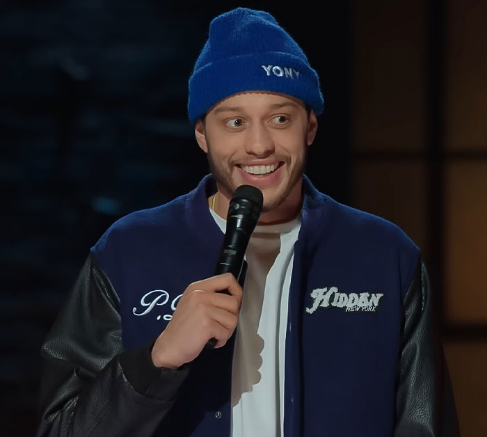 Comedian Pete Davidson on stage holding a microphone, addressing the scrutiny of size of his manhood double standard.