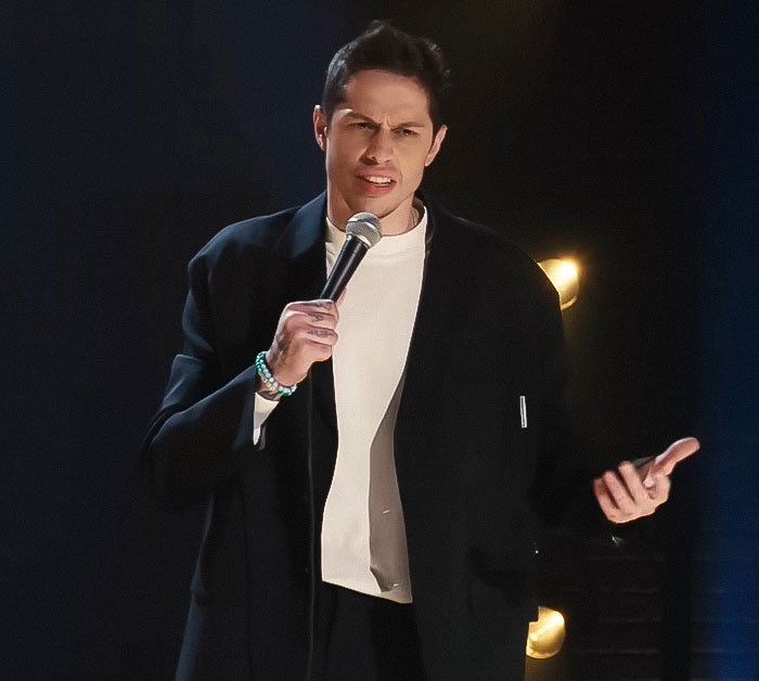 Pete Davidson performing stand-up comedy, addressing the scrutiny and double standard of size of his manhood on stage.