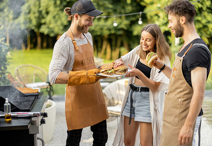 Three neighbors enjoying a backyard barbecue, sharing burgers and smiling on a sunny day outdoors.