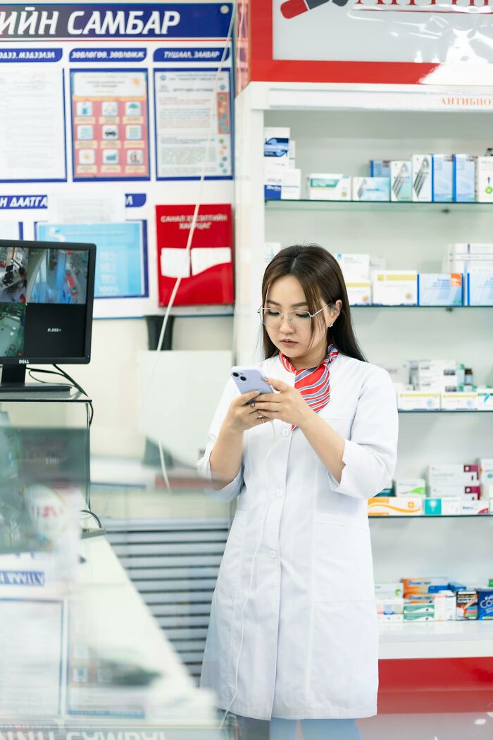 Night shift worker in a pharmacy wearing a white coat and glasses, using a smartphone during work hours.