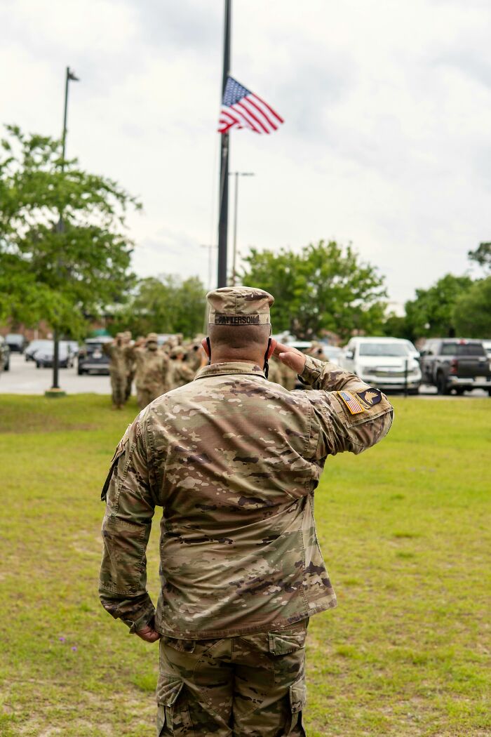 Soldier in camouflage uniform saluting near American flag, illustrating performative behaviors in society discussions.