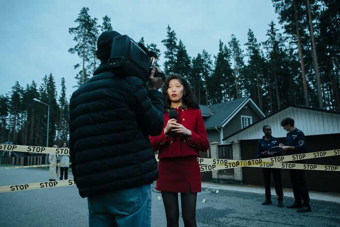 Female reporter in a red outfit filming a true crime story near police tape and officers at a residential crime scene.