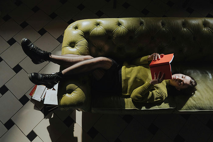 Young woman lying on green couch holding red book, wearing striped tights and black boots in dimly lit room.