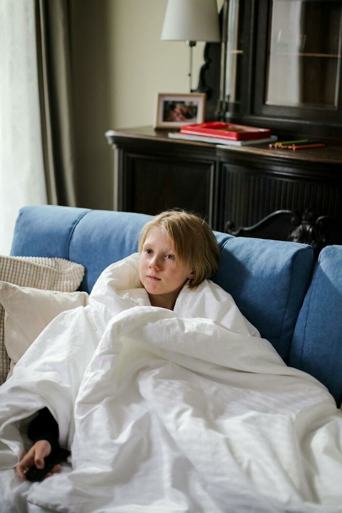 Child wrapped in a white blanket sitting on a blue couch in a cozy room, reflecting on heartwarming secrets.