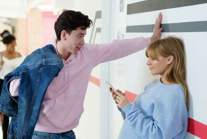 Young man using smooth pickup lines while leaning against a wall and talking to a woman scrolling on her phone.