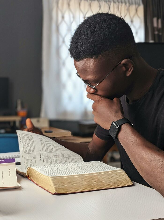 Young man wearing glasses reads a large book at a desk, reflecting on performative behaviors in society.