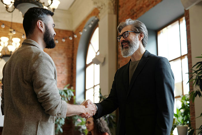 Young man nervously shaking hands with older man before meeting his girlfriend&rsquo;s parents during a tense lunch.