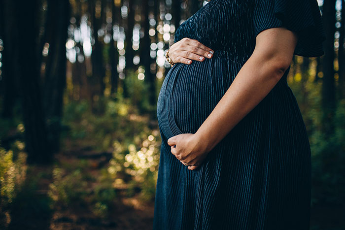 Pregnant woman gently holding belly outdoors in forest, related to woman correcting mother-in-law about baby&rsquo;s name.