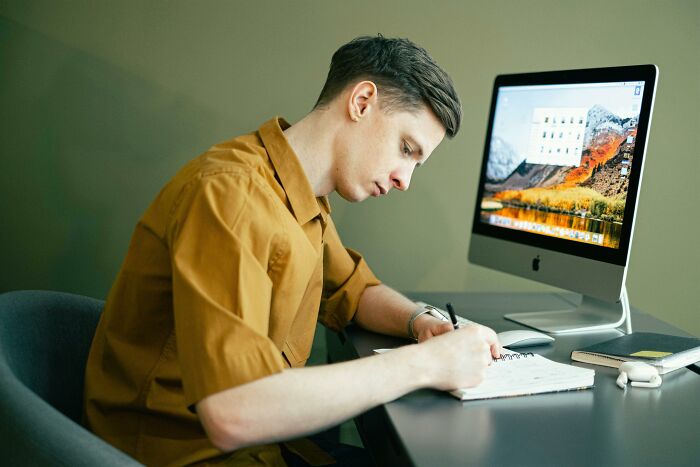 Young man studying geography and business questions, writing notes at desk with computer in a focused setting Young man studying geography and business questions, writing notes at desk with computer in a focused setting