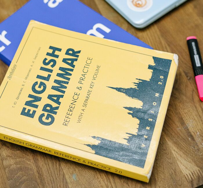 Yellow English grammar book on a wooden table next to a laptop and a black and pink highlighter.