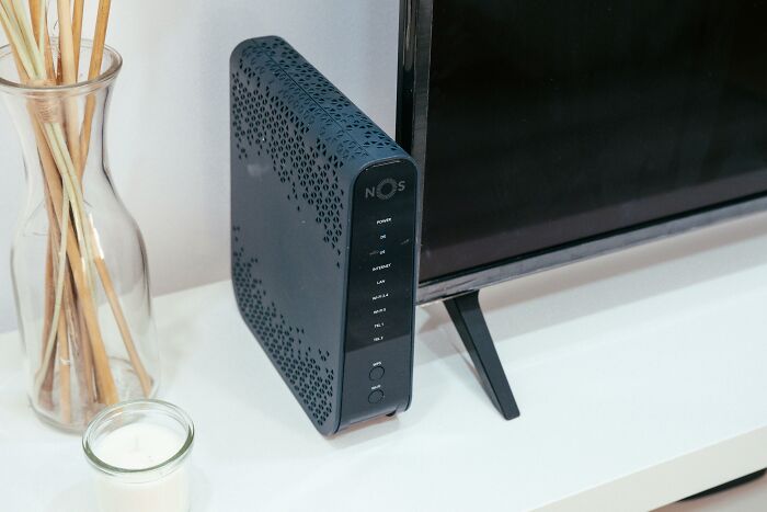 Black modem router next to TV on a white surface with a glass vase and scented candle, illustrating safe things people freak out about.