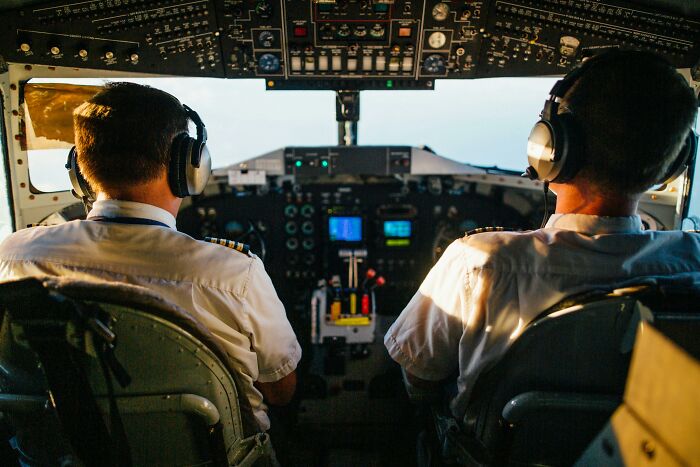 Two pilots in uniform wearing headsets inside a cockpit, focused on controls and instruments ahead.