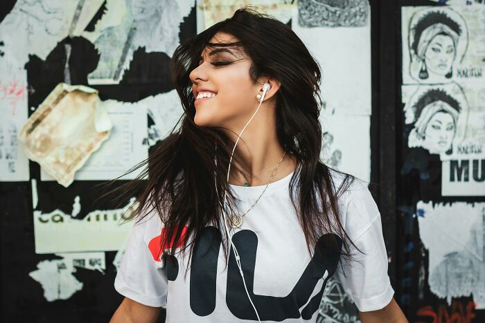 Young woman enjoying music with earphones, smiling and dancing against an urban wall, symbolizing misheard lyrics culture.