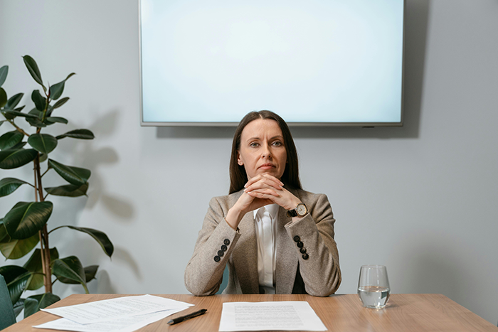 Worker disciplined in a formal office setting, sitting at a desk with documents and glass of water, looking serious.