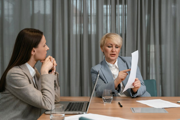 Boss in gray suit pointing at document while employee in beige blazer listens during tense office meeting