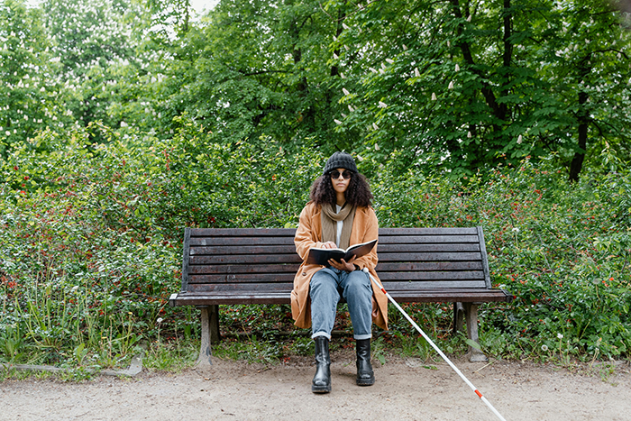 Woman with visual impairment sitting on a bench outdoors using a white blind cane and reading a book surrounded by greenery