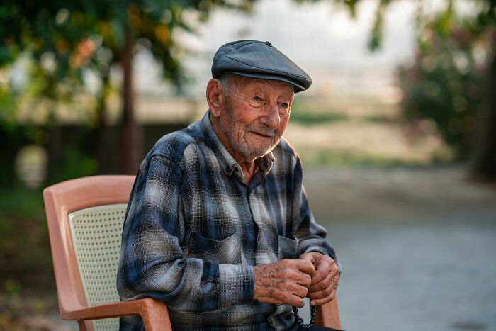 Elderly man wearing a flat cap and plaid shirt sitting outdoors reflecting on hidden family secrets discovered after a loved one passed.