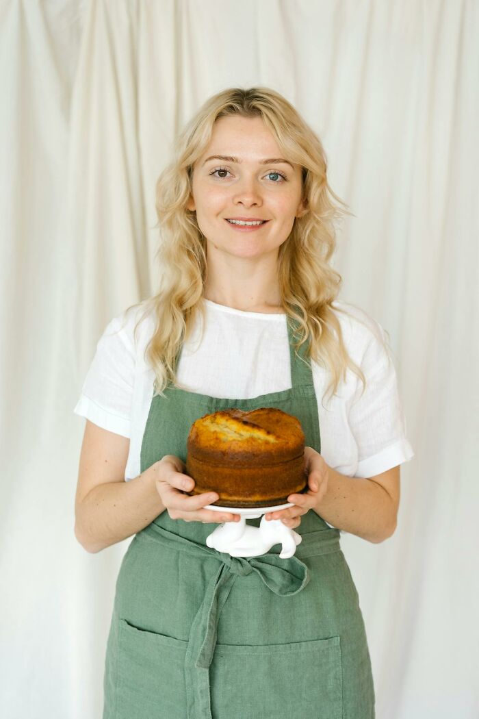 Young woman in green apron smiling and holding a homemade cake, representing heartwarming and harmless secrets concept.