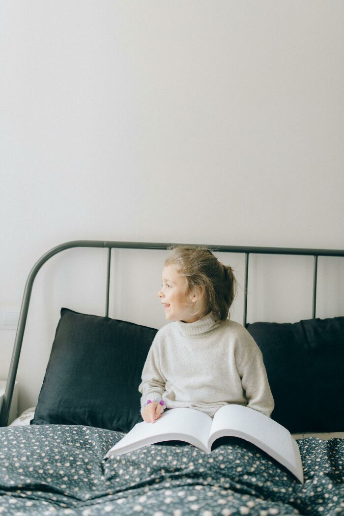 Young girl sitting on bed with an open book, smiling and looking away, related to creepy and terrifying imaginary friends.