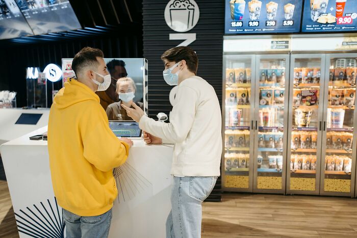 Two men wearing masks discussing profession secrets while standing at a concession stand in a movie theater.