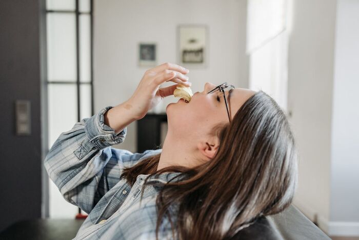 Young woman in glasses eating a pastry indoors, capturing a heartwarming and harmless secret moment.