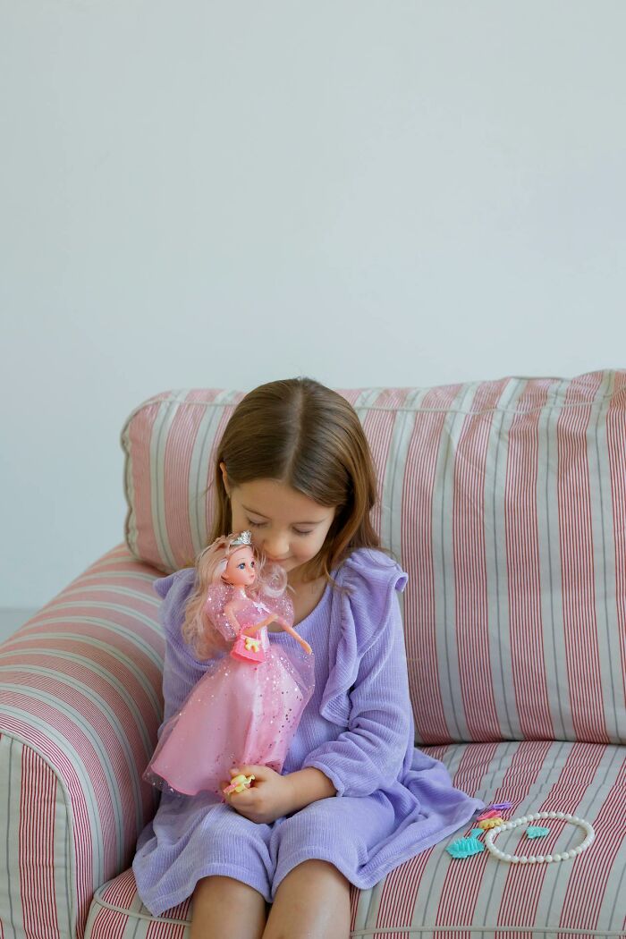 Young girl in purple dress sitting on striped couch holding doll, evoking creepy and terrifying experiences with imaginary friends
