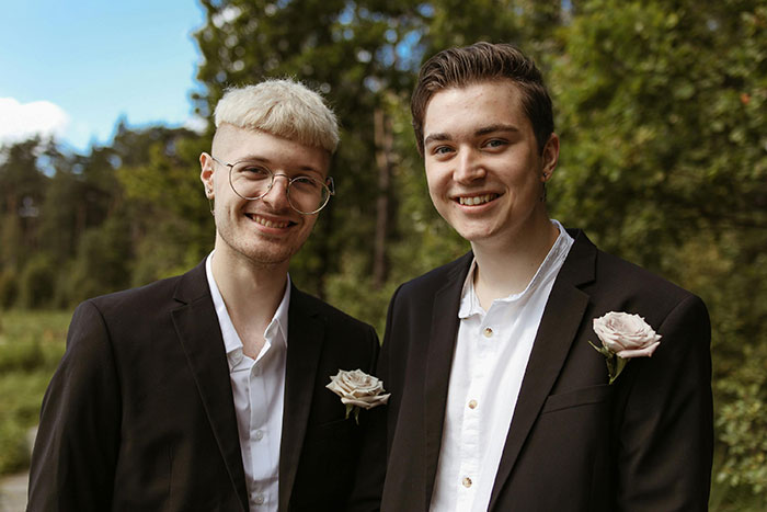 Two young men in suits with boutonnières smiling outdoors, representing a man urging gay brother to come out. Two young men in suits with boutonnières smiling outdoors, representing a man urging gay brother to come out.