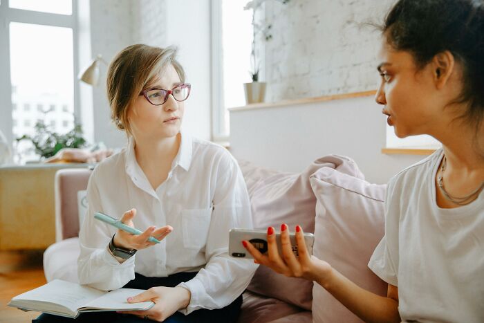 Two women discussing misheard phrases, one with glasses holding a notebook, in a bright modern living room.