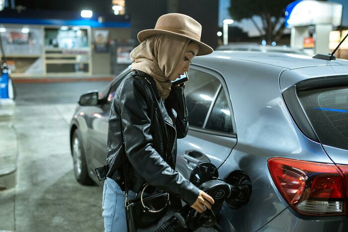Woman in a hat and leather jacket fueling a car at night, illustrating totally safe things that cause people to freak out.