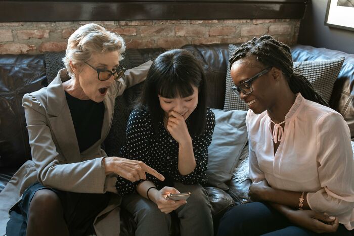 Three women sitting on a couch, smiling and reacting to a phone, sharing stories about picking the wrong line at the grocery store.