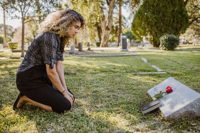 Young woman kneeling at a gravestone in a cemetery, reflecting quietly, conveying heartwarming and harmless secrets.