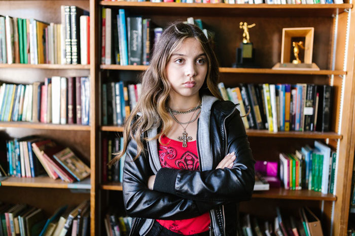 Teen girl with crossed arms looks upset in front of a bookshelf, reflecting challenges of winning over fiancee&rsquo;s kids.
