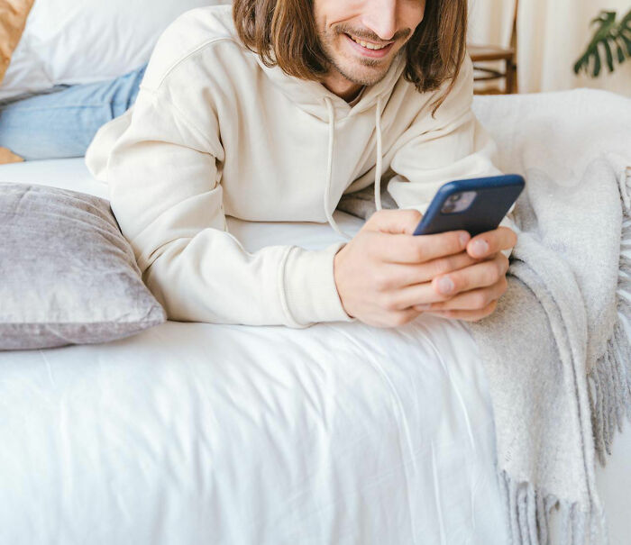 Young man in a cream hoodie smiling while lying on bed and using smartphone, reflecting signs of talking to a psychopath online.