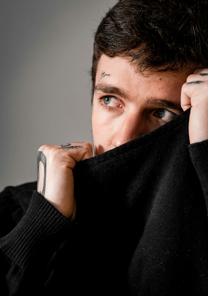 Young man with tattoos covering his face and hands, hiding behind a black sweater, evoking paranormal encounter vibes.